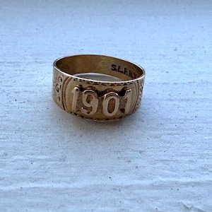 May include: A gold-toned metal ring featuring the embossed number 1901 on the front surface. The band displays decorative scrollwork patterns on the sides and includes the engraved text S.L.F.N.Y. on the interior surface of the ring band.