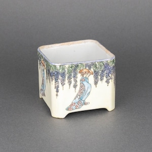 May include: A square white ceramic planter with a floral design featuring a woman in a blue and white kimono standing under a purple wisteria vine. The planter has a light brown rim and four feet.