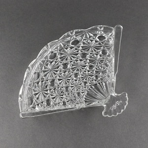 May include: A clear glass napkin holder with a diamond pattern. The holder has a fan-shaped design and a small, decorative handle.