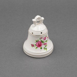 Puede incluir: Salero de cerámica blanca en forma de campana con un diseño floral de rosas rosas y verdes. El salero tiene un borde dorado y un asa en la parte superior. La campana tiene varios agujeros.