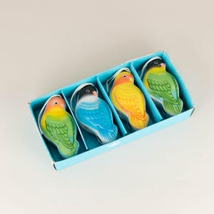 May include: Four colourful bird-shaped candles in a blue box. The candles are yellow, green, blue, and green with yellow accents.