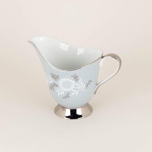May include: A light blue creamer with a silver floral design and a silver rim. The creamer has a silver base.