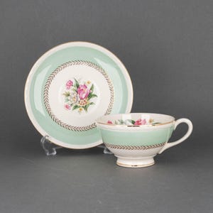 May include: A vintage teacup and saucer set with a floral design. The cup and saucer are white with a green rim and a gold trim. The floral design features pink and white flowers.