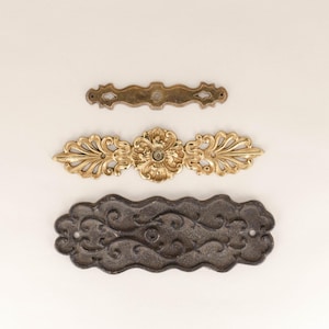 Puede incluir: Tres tiradores de cajón decorativos de metal. El tirador superior es un tirador simple, plano y de latón. El tirador del medio es un tirador más ornamentado, de color dorado, con un diseño floral. El tirador inferior es un tirador ondulado de metal negro con un diseño floral.