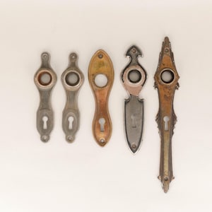 May include: Five antique door escutcheons in various shapes and finishes. The escutcheons are made of metal and feature keyhole openings. Colors include silver, bronze, and gold, with signs of age and wear.