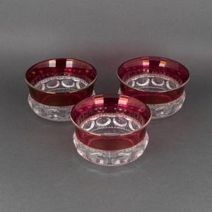 Puede incluir: Tres cuencos de vidrio transparente con una banda roja rubí y ribete dorado. Los cuencos tienen un patrón decorativo en la base. Los cuencos están dispuestos sobre una superficie gris.