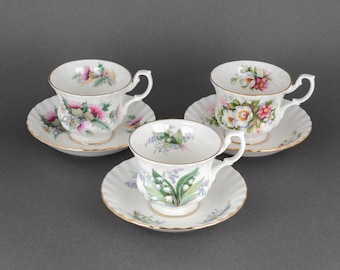 May include: Three white teacups and saucers with floral designs. The cups have a gold trim and the saucers have a scalloped edge. The floral designs include thistles, lilies, and roses.