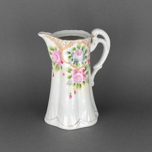 Peut inclure: Un pichet en porcelaine blanche avec un motif floral. Le pichet a une base large et un bec étroit. Le motif floral présente des roses roses et blanches avec des feuilles vertes.
