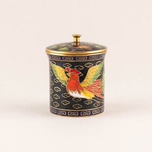 Puede incluir: Un pequeño tarro redondo de cloisonné negro y dorado con tapa. El tarro está decorado con un diseño de ave fénix colorido y un borde azul y dorado.