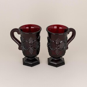 Puede incluir: Dos tazas de vidrio rojo oscuro con base y un patrón decorativo. Las tazas tienen un asa y están hechas de vidrio rojo rubí.
