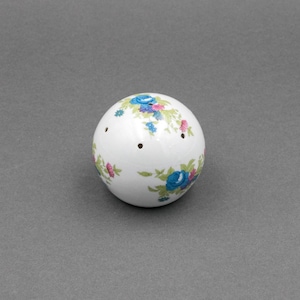 May include: A white ceramic ball-shaped salt shaker with a floral pattern in blue, pink, and green. The shaker has three small holes and is designed for dispensing salt or other seasonings. The floral design adds a decorative touch.