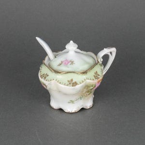 May include: A white porcelain sugar bowl with a lid and a small spoon. The bowl is decorated with a green and gold floral pattern.