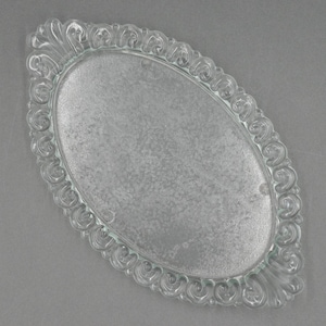 Puede incluir: Bandeja ovalada de vidrio transparente con superficie texturizada. La bandeja tiene un borde decorativo y ornamentado con un diseño de volutas repetidas. La bandeja está vacía y se encuentra sobre una superficie gris.