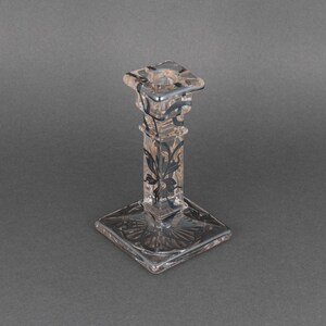 May include: A clear glass candlestick with a square base and a square top. The candlestick has a black floral design etched into the glass.