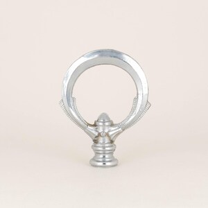 May include: A silver metal finial with a circular ring at the top and a decorative, pointed finial at the bottom.