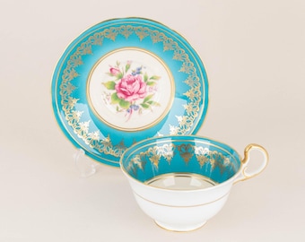 May include: A white teacup and saucer with a turquoise blue rim and gold trim. The saucer has a floral design in the centre.
