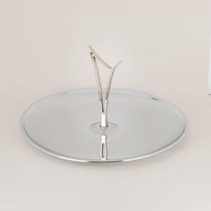 May include: A silver-coloured serving tray with a central handle. The round tray has a polished, reflective surface and a decorative handle that curves upwards. The handle is a stylized V-shape. Suitable for serving cakes, pastries, or other treats.