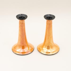 May include: A pair of matching candlestick holders featuring a lustrous, iridescent orange finish that reflects light. Each holder has a wide, flared base that tapers upward to a narrow neck, topped with a circular, solid black rim for candles.