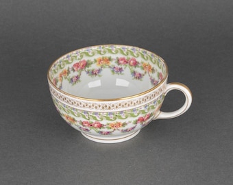 May include: A white porcelain teacup with a floral pattern in shades of pink, green, and yellow. The cup has a gold rim and a delicate floral design.