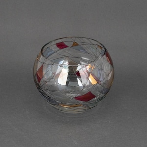 Puede incluir: Un portavelas de vidrio transparente con un patrón geométrico de rojo, dorado y azul. El portavelas es redondo y tiene una abertura ancha.