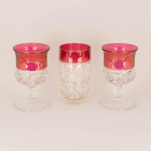 May include: Three clear drinking glasses with a ruby red rim. Two glasses have a stemmed base with a cut glass design, and the third is a straight-sided tumbler. The glasses are empty and sit on a white surface.