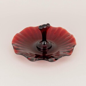 Puede incluir: Una fuente de vidrio rojo rubí con un asa central. La fuente tiene un borde festoneado y una textura acanalada, creando una estética vintage. El asa es de color oscuro y sólido, y la fuente se utiliza probablemente para servir postres o pequeños aperitivos.