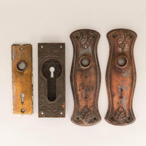 以下が含まれることがあります： 鍵穴のあるアンティークなドア金具プレート4枚。2枚は装飾が施され、銅仕上げ。1枚は長方形で黒仕上げ。最後の1枚は長方形で金仕上げ。