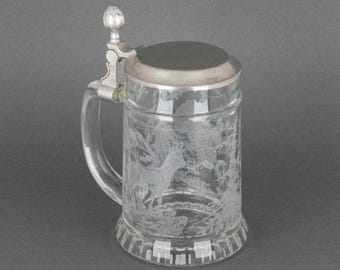 May include: Clear glass beer stein with a silver-colored lid and handle. The glass is etched with a forest scene featuring a deer. The lid is hinged and has a decorative knob. The stein has a classic, traditional design.