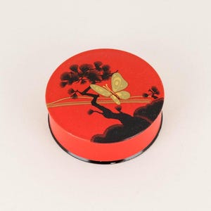 May include: A round red trinket box with a black and gold design of a pine tree and a butterfly. The box has a black rim.