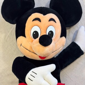 May include: A plush Mickey Mouse doll with black ears, a black head, and a tan face. The doll has white gloves, a red pair of shorts, and a yellow button. The doll is smiling and has its arm raised.
