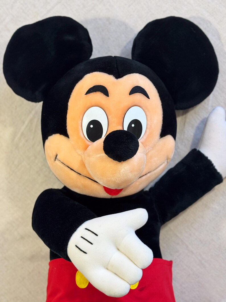 May include: A plush Mickey Mouse doll with black ears, a black head, and a tan face. The doll has white gloves, a red pair of shorts, and a yellow button. The doll is smiling and has its arm raised.