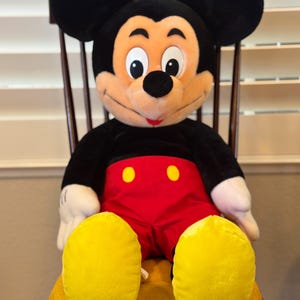 Könnte beinhalten: Eine große Mickey Mouse Plüschpuppe, die auf einem Holzstuhl sitzt. Mickey trägt rote Shorts mit gelben Knöpfen, weiße Handschuhe und gelbe Schuhe. Seine schwarzen Ohren und sein schwarzer Körper sind sichtbar. Die Puppe hat einen lächelnden Gesichtsausdruck.