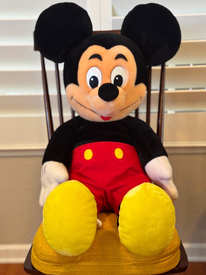 May include: A large plush Mickey Mouse doll, seated on a wooden chair. Mickey is wearing red shorts with yellow buttons, white gloves, and yellow shoes. His black ears and black body are visible. The doll has a smiling expression.