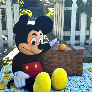 May include: A plush Mickey Mouse character sits on a picnic blanket with a brown wicker basket and a small orange pumpkin. The blanket has a yellow border with blue and white designs. Mickey is wearing a black body suit, red shorts, and yellow shoes.