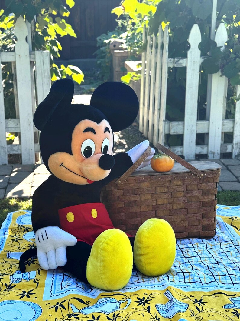 May include: A plush Mickey Mouse character sits on a picnic blanket with a brown wicker basket and a small orange pumpkin. The blanket has a yellow border with blue and white designs. Mickey is wearing a black body suit, red shorts, and yellow shoes.