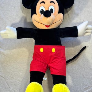 May include: A large plush Mickey Mouse toy. The iconic character has a black body, red shorts with yellow buttons, yellow shoes, and white gloves. Mickey's arms are outstretched, and his face is smiling.