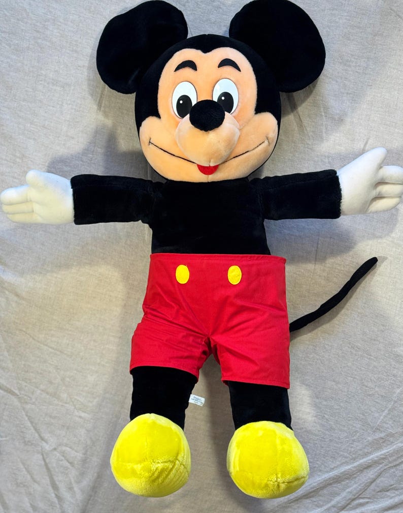 May include: A large plush Mickey Mouse toy. The iconic character has a black body, red shorts with yellow buttons, yellow shoes, and white gloves. Mickey's arms are outstretched, and his face is smiling.