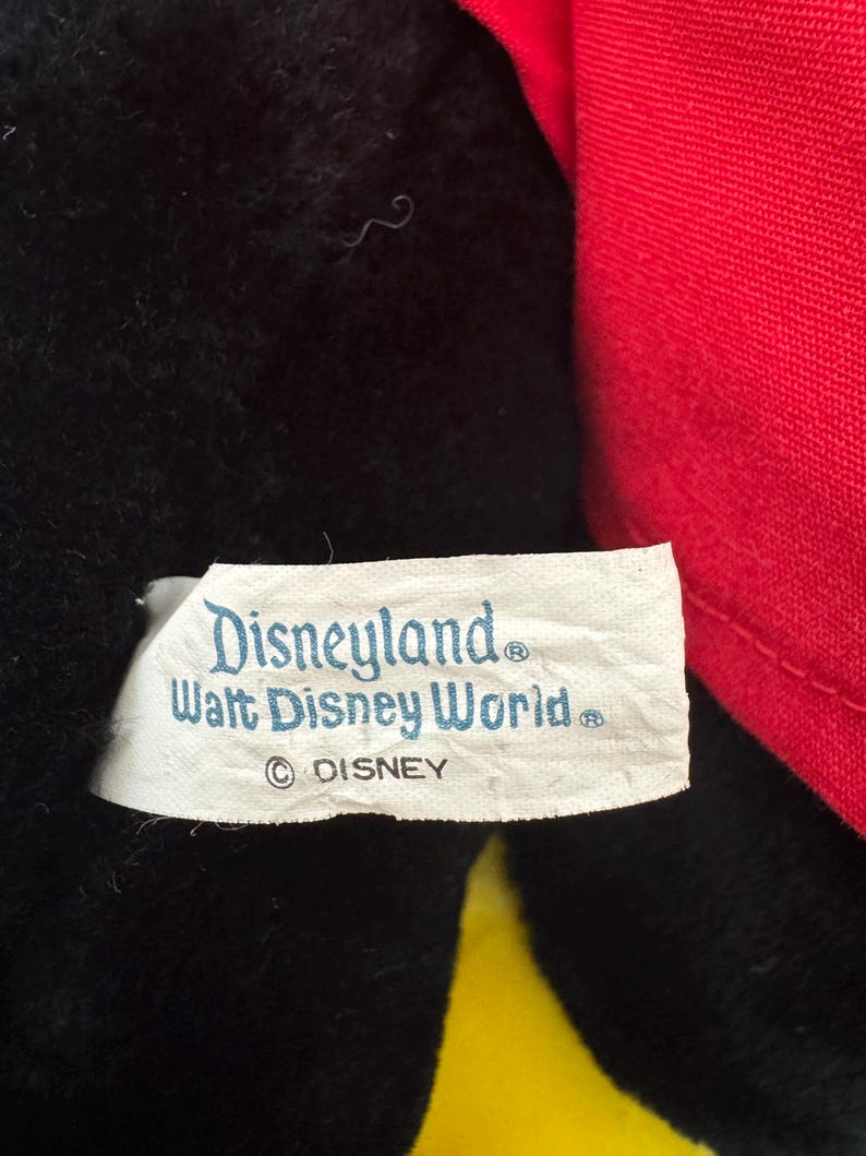 May include: Close-up of a plush toy with black fur and a red fabric element. A white tag displays the text "Disneyland Walt Disney World" and the Disney copyright symbol.
