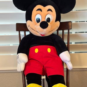 May include: A plush Mickey Mouse doll, featuring a black body, large round ears, and a smiling face. He wears red shorts with yellow buttons, white gloves, and yellow shoes. The doll is seated on a wooden chair.