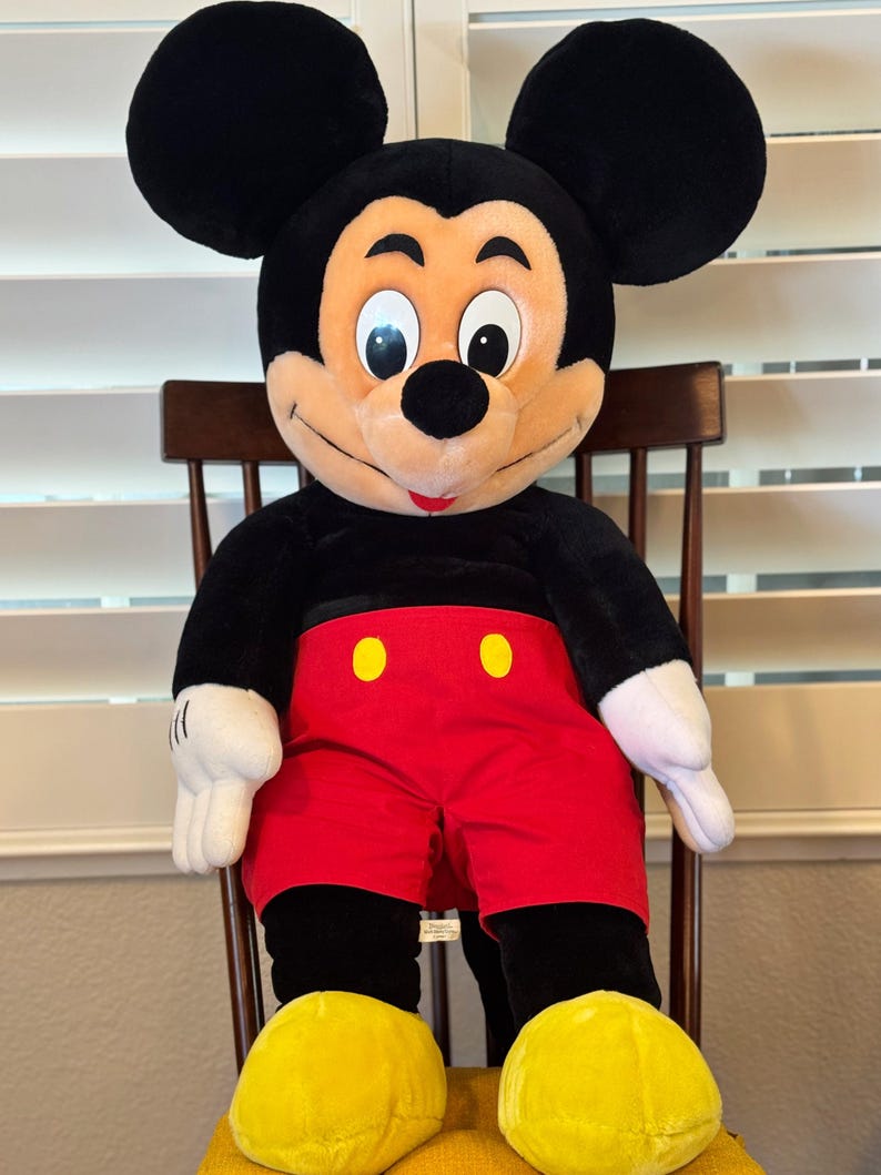 May include: A plush Mickey Mouse doll, featuring a black body, large round ears, and a smiling face. He wears red shorts with yellow buttons, white gloves, and yellow shoes. The doll is seated on a wooden chair.