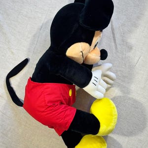 May include: A plush Mickey Mouse toy, featuring a black body, ears, and tail, with a tan face and white gloves. Mickey wears red shorts with yellow buttons and large yellow shoes. The toy is lying on a light gray surface.