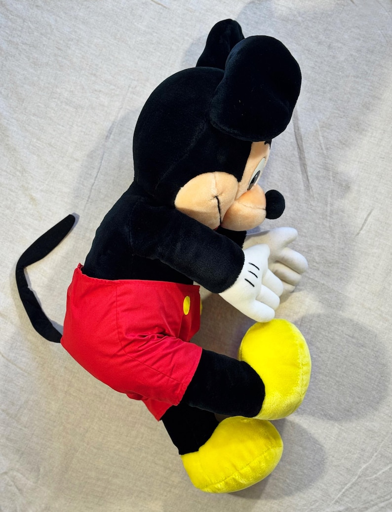May include: A plush Mickey Mouse toy, featuring a black body, ears, and tail, with a tan face and white gloves. Mickey wears red shorts with yellow buttons and large yellow shoes. The toy is lying on a light gray surface.