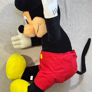 May include: A plush Mickey Mouse doll in a seated position. The doll features a black head and body, red shorts with a yellow button, yellow shoes, and white gloves. The doll's right hand is raised.