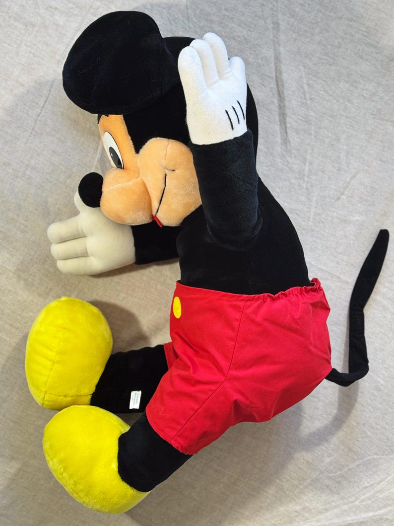 May include: A plush Mickey Mouse doll in a seated position. The doll features a black head and body, red shorts with a yellow button, yellow shoes, and white gloves. The doll's right hand is raised.