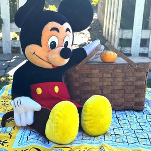 May include: A large Mickey Mouse plush toy with black ears, a red shorts, and yellow shoes sits on a picnic blanket. A woven picnic basket and a small orange fruit are nearby. The blanket has a yellow and blue pattern.
