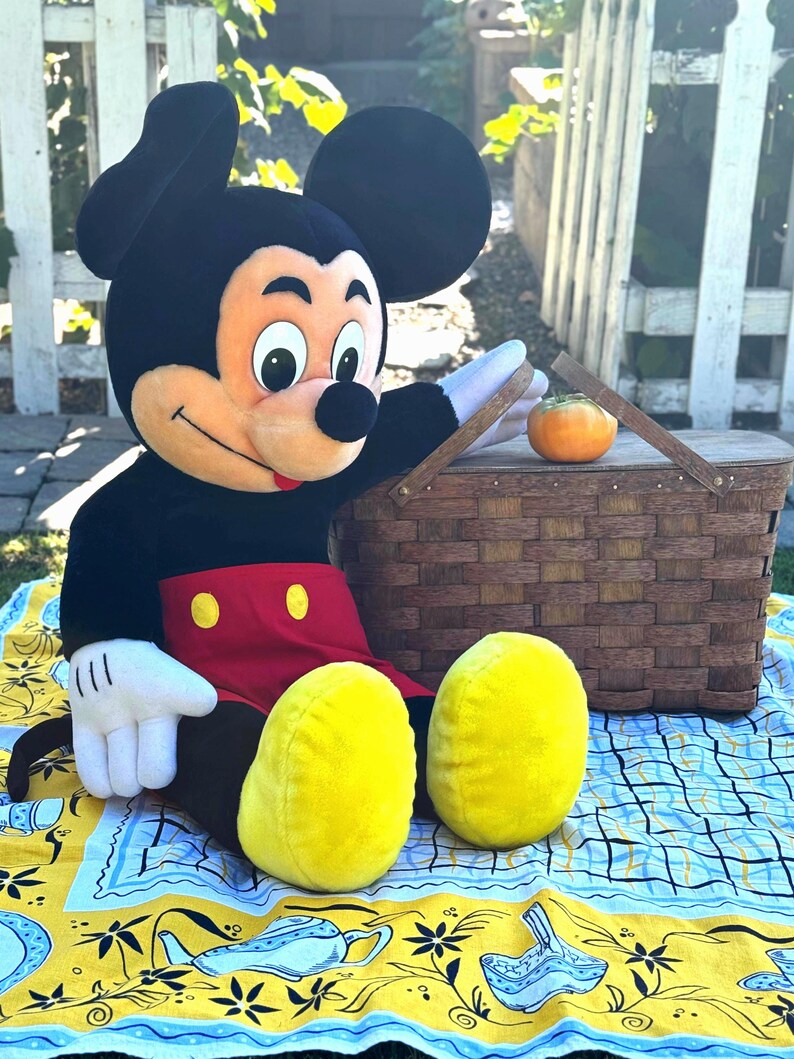 May include: A large Mickey Mouse plush toy with black ears, a red shorts, and yellow shoes sits on a picnic blanket. A woven picnic basket and a small orange fruit are nearby. The blanket has a yellow and blue pattern.