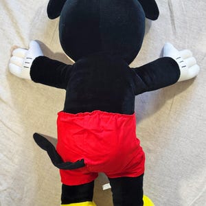 May include: A plush Mickey Mouse toy, viewed from the back. The toy features a black body, red shorts, white gloves, and yellow shoes. The arms are outstretched, and the tail is visible.