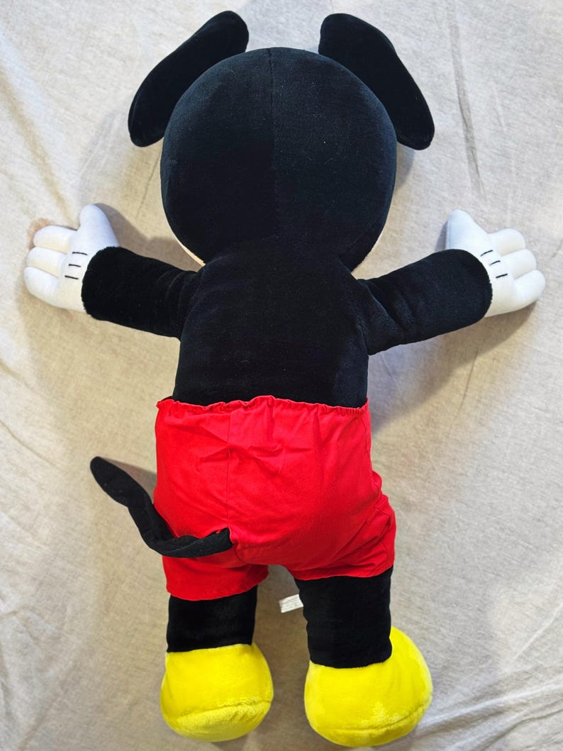 May include: A plush Mickey Mouse toy, viewed from the back. The toy features a black body, red shorts, white gloves, and yellow shoes. The arms are outstretched, and the tail is visible.