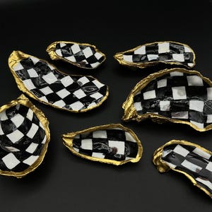 May include: Several oyster shell dishes with a black and white checkered pattern inside and gold-painted edges. The shells vary in size and shape, arranged on a black surface.