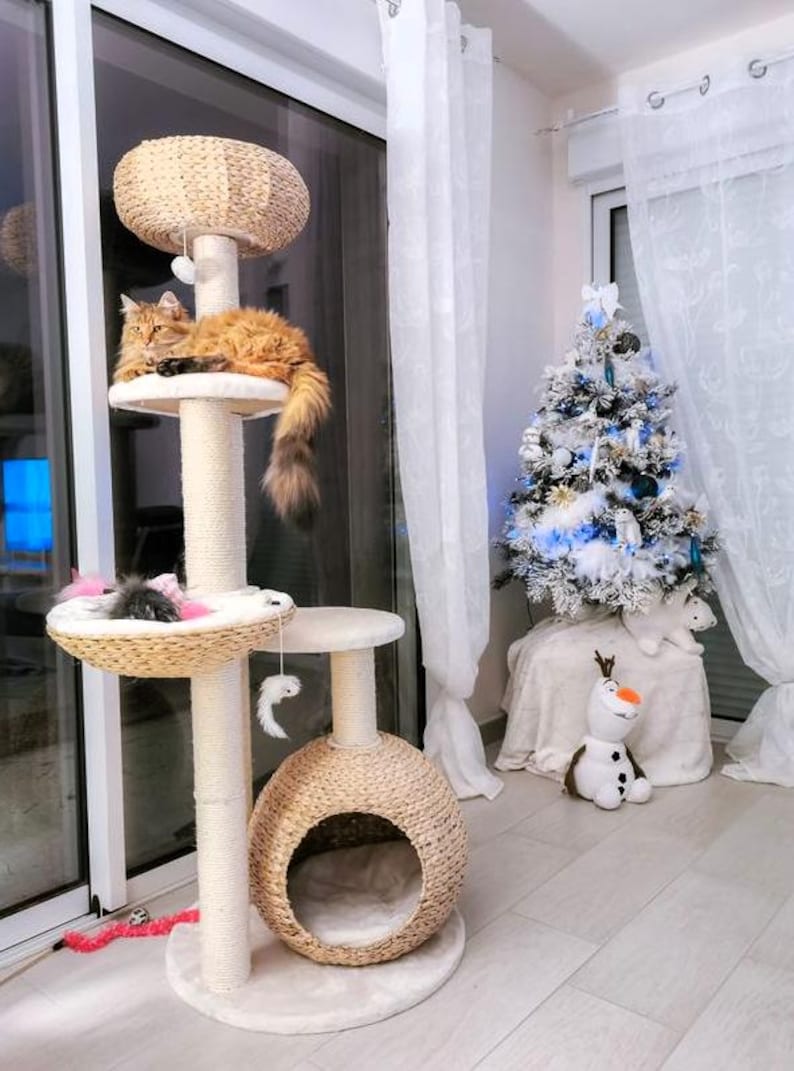May include: A beige cat tree with a brown cat lounging on the top platform. The cat tree has a round bed at the bottom, a round platform in the middle, and a round platform at the top. The cat tree is in a room with white curtains and a small white Christmas tree decorated with blue and white ornaments.